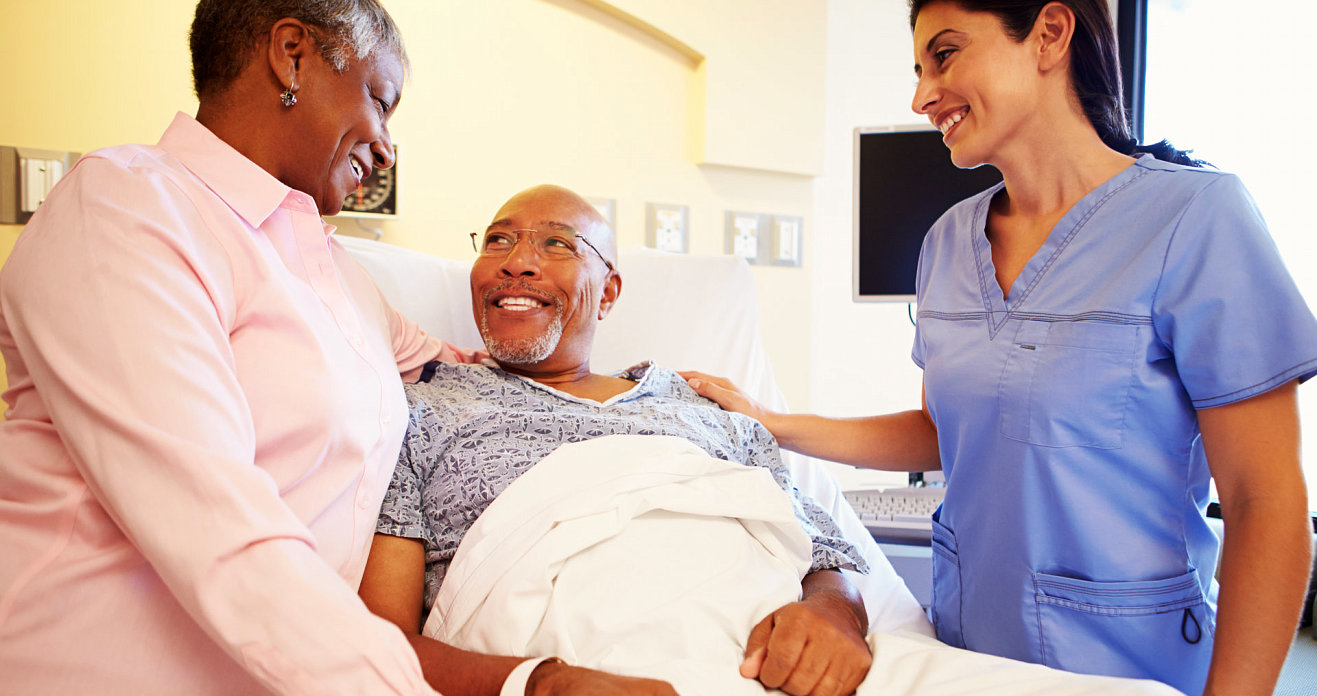 a senior man in hospital bed smiling with his wife and caregiver